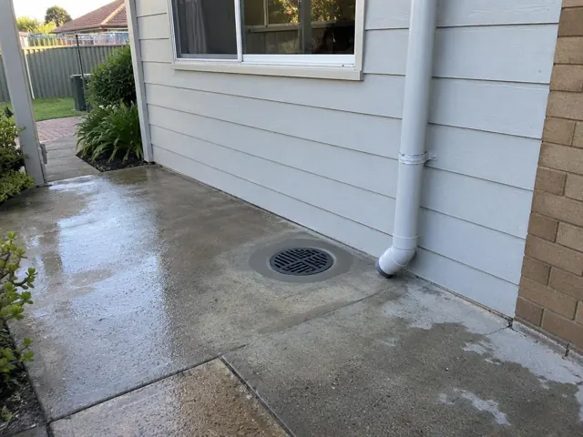 Outdoor drain grate set in wet concrete paving beside a weatherboard house in Matraville